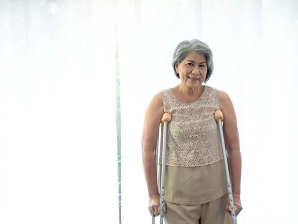 Asian senior woman white hair standing with crutches near curtain with copy space, smile and looking at camera. Elderly lady patient using crutches walker. Strong health, medical care concepts.