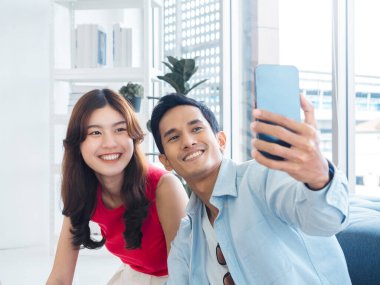 Happy handsome Asian man and young beautiful woman selfie while sitting near the glass window at white living room in high building. Cheerful couple taking portrait selfie with smart mobile phone.