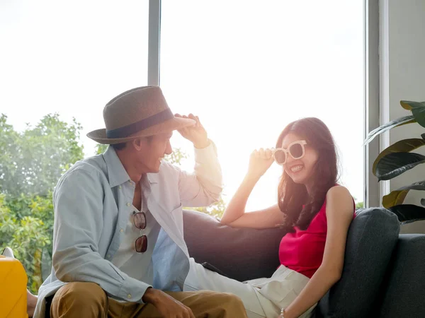 Happy Asian couple lover ready to trip, young woman and man in colorful casual wearing beach hat and sunglasses, smiling together, sit on couch in living room on sunset, travel on summer holiday.