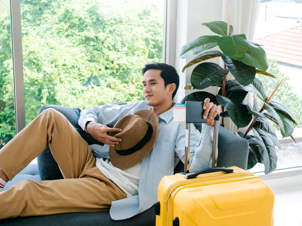 A man with yellow luggage. Handsome Asian male in denim shirt sitting and waiting alone while holding hat and yellow suitcase near the glass window. Summer holiday travel vacation concept.