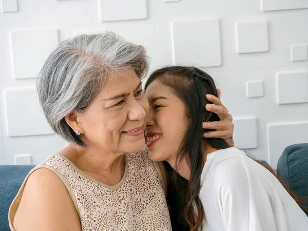 Close up happy Asian senior, mother white hair embracing her beautiful daughter while she kiss her cheek with love, care and smile while sit on sofa on white background in living room at home.