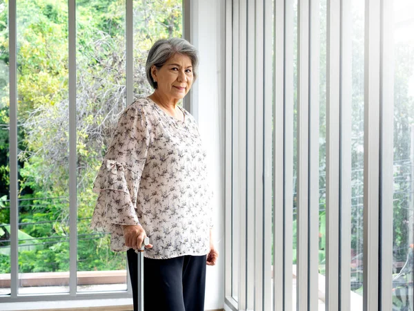 Asian senior woman, white hair standing with a cane near the glass window, indoors. Elderly lady patient using walking cane. Strong health, medical care and life insurance concepts.
