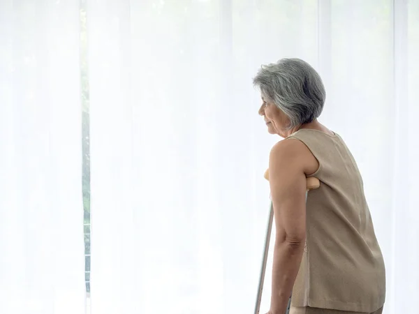 Asian senior woman white hair standing with walking crutches near curtain looking out glass window with copy space. Elderly lady patient using crutches walker. Strong health, medical care concepts.