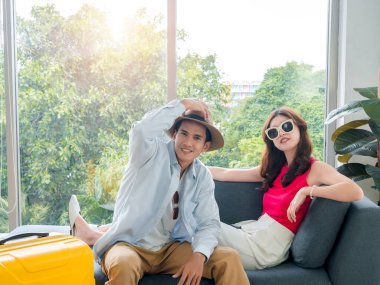 Happy Asian couple lover ready to trip, young woman and man in colorful casual wearing beach hat and sunglasses, smiling together, sit on couch with baggage in living room, travel on summer holiday.