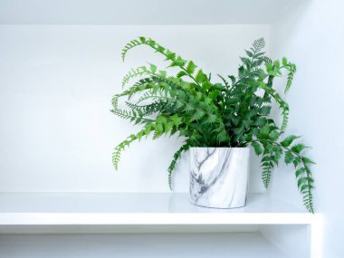 White modern ceramic plant pot with artificial plant with green leaves on white shelf with copy space.