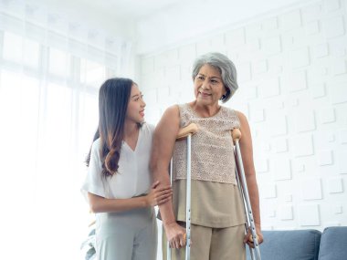 Elderly woman trying to walk on crutches standing held and supported in arms by young Asian female carefully in recovery room, helping old women, health care, senior therapy patient at home concept.