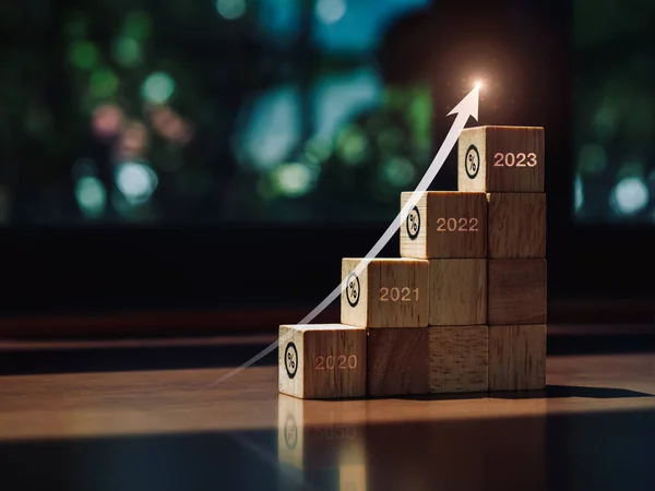 Shining rise up arrow on wood blocks chart steps with percentage icons from year 2020 to 2023 on wooden desk, business investment process and success, economic improvement, and growth trends concept.