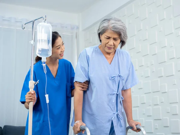 Elderly Asian woman patient trying to walk on walking frame held and carefully supported in arms by caregiver, young polite female assistant nurse in blue suit in white room, senior care concept.