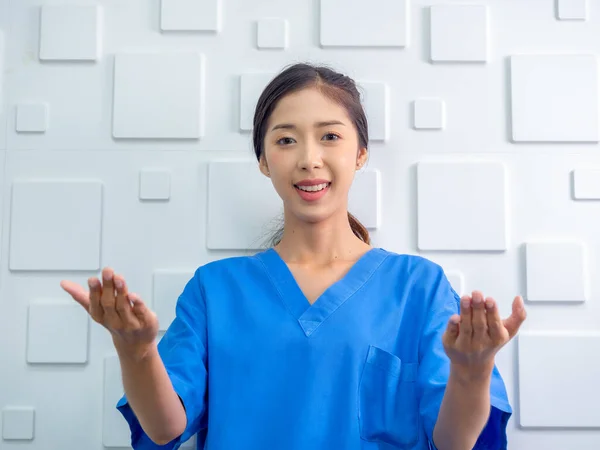 Portrait of friendly, cheerful, smile confident Asian woman doctor or nurse doing explanatory gesture with her hands on white background, looking at camera. Healthcare professional in blue scrubs.