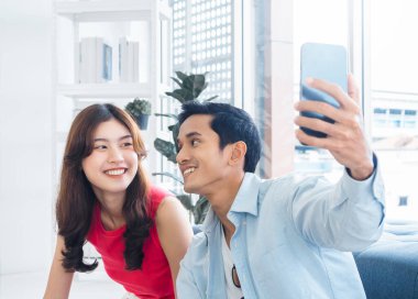 Happy handsome Asian man and young beautiful woman selfie while sitting near the glass window at white living room in high building. Cheerful couple taking portrait selfie with smart mobile phone.