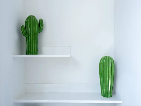 Two green ceramic cactus doll model decoration on two steps of white shelf. Home decor with fake plant object.