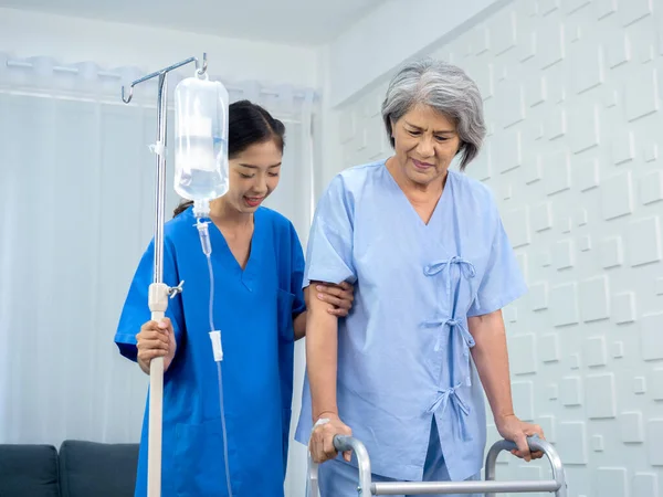 Elderly Asian woman patient trying to walk on walking frame held and carefully supported in arms by caregiver, young polite female assistant nurse in blue suit in white room, senior care concept.