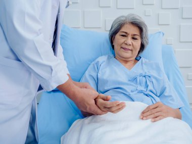 Hastanede kalp atışlarını kontrol etmek için beyaz laboratuvar önlüklü doktor el ele tutuşurken mutlu Asyalı yaşlı hasta kadın yatakta gülümsüyor. Sağlık hizmetleri kontrolü kavramı.