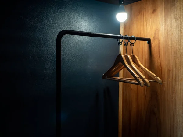 Three empty wooden clothes hangers without shirts or dress hanging on a black cloth rack with light bulb in the wood wardrobe on dark blue background preparing for the guest who staying in hotel room.