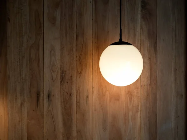 Round hanging lamp, full moon shape with the light bulb inside shining in the wood wall background in the dark room with copy space.