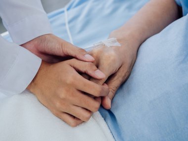 Beyaz takım elbiseli yakın plan kadın doktor, yaşlı bir kadının elini tutuyor. Açık mavi elbiseli, hastane yatağında salin hortumlu bir kadın. Hasta kavramını destekliyor..