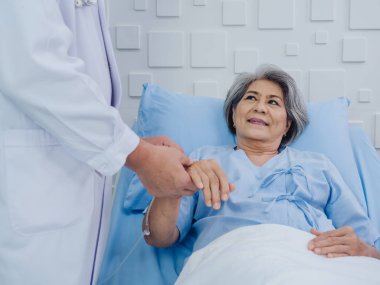 Mutlu bir gülümseme, beyaz takım elbiseli erkek doktor elini tutarken ve hastane odasında damardan sıvı verirken, açık mavi elbiseli, yakışıklı, yaşlı bir kadın..