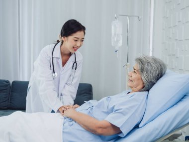 Tuz solüsyonuyla yatakta uzanan açık mavi elbiseli mutlu Asyalı yaşlı kadın hasta steteskoplu ve hastane odasında el ele tutuşan beyaz takım elbiseli genç bir doktor tarafından teşvik ediliyordu..