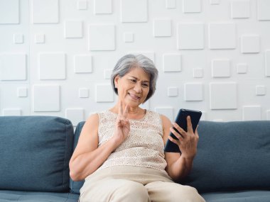 Mutlu Asyalı son sınıf öğrencisi kadın beyaz odada el ele dijital tablet ekranda zafer işareti yapıyor. Yaşlı bir kadın evde ailesiyle görüntülü görüşme yapıyor. Teknolojisi olan yaşlı insanlar..