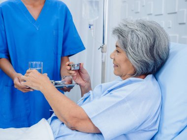 Asyalı yaşlı bir hasta hastane odasında mavi önlüklü genç hemşireden su bardağı ve hap alıyor, günlük ilaç veya vitamin takviyesi, kıdemli sağlık ve tıbbi destek alıyor..