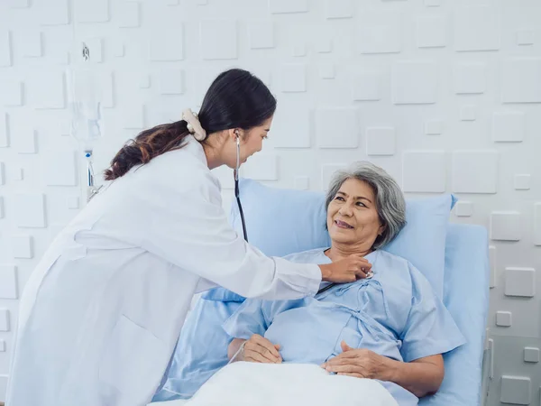 Happy smile Asian elderly old female patient in light blue dress lying on bed while beautiful young woman doctor in white suit using stethoscope to examining, listen to her heartbeat in hospital room.