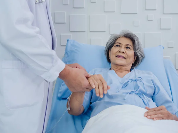 Happy smile beautiful Asian elderly old woman patient in light blue dress lying on bed while male doctor in white suit holding her hand and giving intravenous fluid on hand in the hospital room.