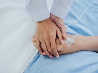 Beyaz takım elbiseli yakın plan kadın doktor, yaşlı bir kadının elini tutuyor. Açık mavi elbiseli, hastane yatağında salin hortumlu bir kadın. Hasta kavramını destekliyor..