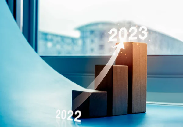 Shining rise up arrow on wood blocks chart steps as a graph from year 2022 to 2023 on blue background with cityscape, business growth process, profit, wealth, trends, economic improvement concepts.