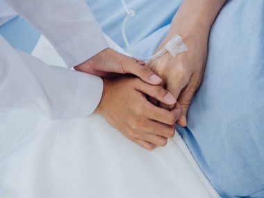 Beyaz takım elbiseli yakın plan kadın doktor, yaşlı bir kadının elini tutuyor. Açık mavi elbiseli, hastane yatağında salin hortumlu bir kadın. Hasta kavramını destekliyor..