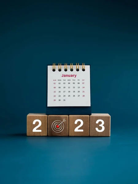 Happy New Year 2023. 2023 year number with target icon on wooden cube blocks and the small white desk calendar on January page on blue background, vertical style. Begin a new business goal to success.