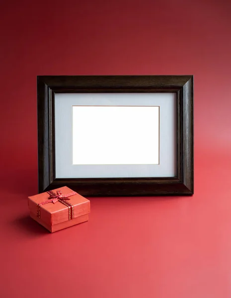 Gift box and photo frame. Template for photo or card with a gift, minimal style. Empty white blank space in vintage wooden photo frame with small red present box on red background, vertical style.