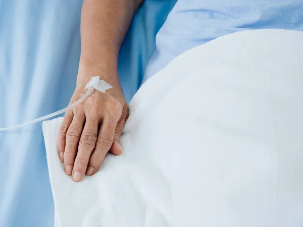 Close up saline solution in senior patients hand in blue gown lying on the bed with white blanket in hospital room with copy space. Elder patient's hand receiving iv saline solution.
