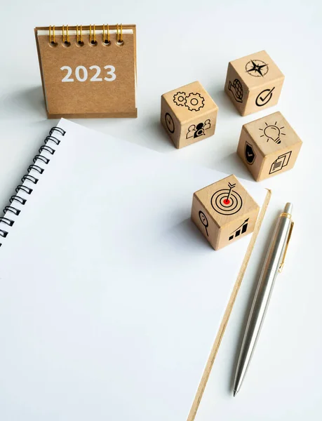 Business process and plan to success icons wooden cube blocks with 2023 small desk calendar year on blank notepad with pen on white background with copy space, vertical style. Happy new year 2023.