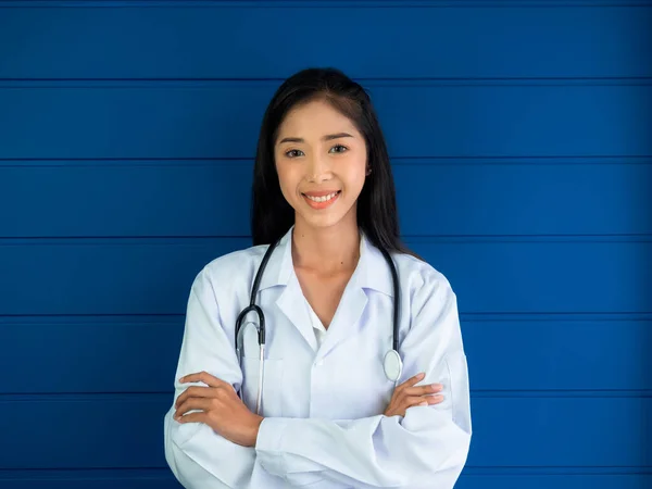 Smiling pretty Asian woman doctor portrait standing on blue wood background in medical office in hospital or clinic. Confident Asian young female doctor with stethoscope, looking at camera.