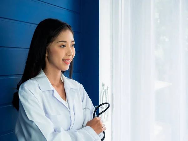 Smiling pretty Asian woman doctor portrait standing on blue wood background near the curtain window in medical office with copy space. Confident Asian young female doctor holding stethoscope.
