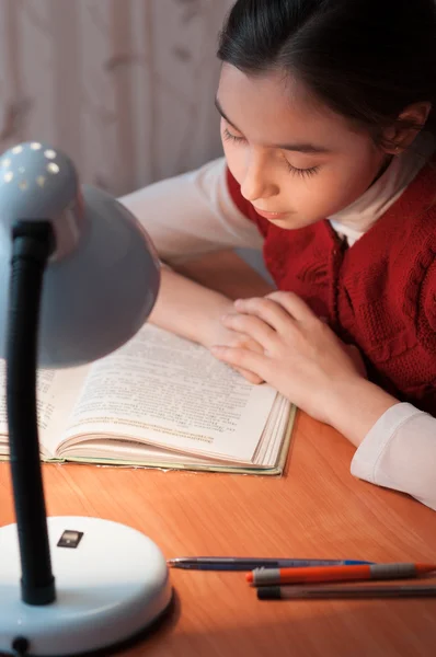 Menina na mesa lendo um livro pela luz da lâmpada — Fotografia de Stock