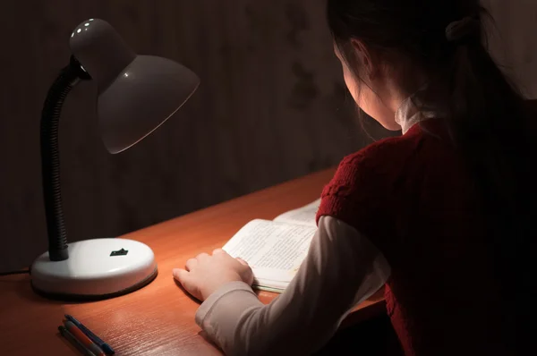 Menina na mesa lendo um livro pela luz da lâmpada — Fotografia de Stock