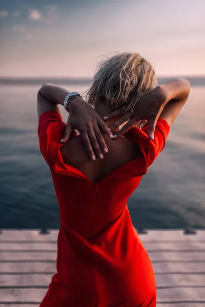 Nehir kenarındaki düğmesiz kırmızı elbiseli bir kadının arka görüntüsü. İskelede atmosferik gün batımı. — Stok fotoğraf