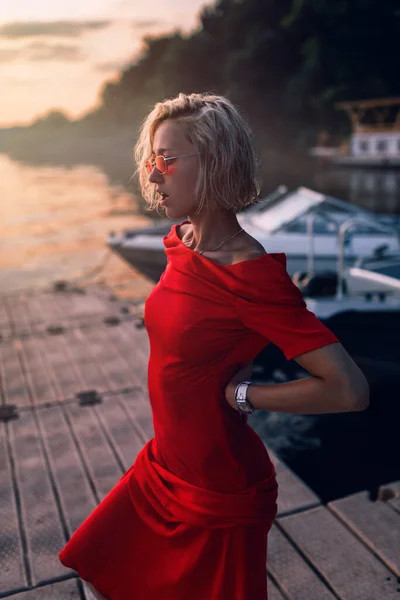 Una mujer rubia con un vestido rojo se para en el muelle. —  Fotos de Stock