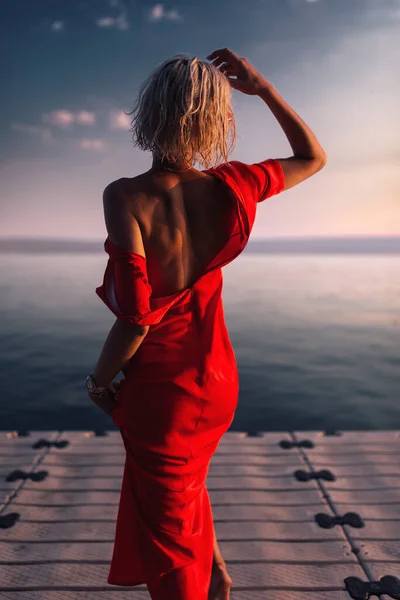 Düğmeleri iliklenmemiş kırmızı elbiseli muhteşem bir sarışın iskelede duruyor.. — Stok fotoğraf