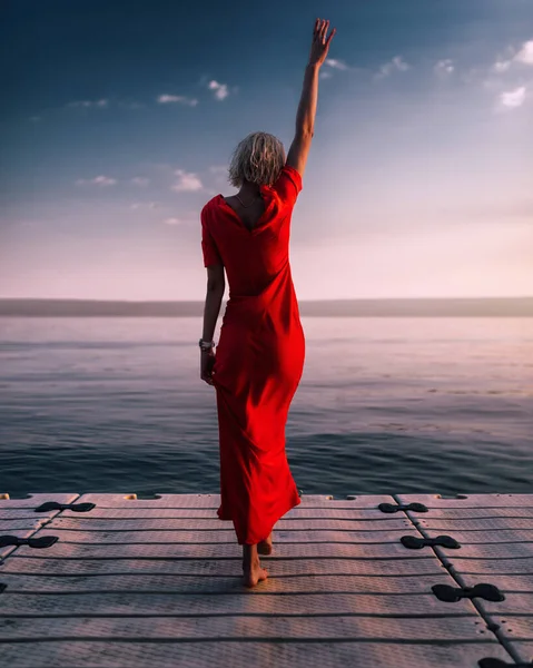 Attraente giovane donna si trova vicino al lago in abito rosso. Tramonto atmosferico sul molo. — Foto Stock