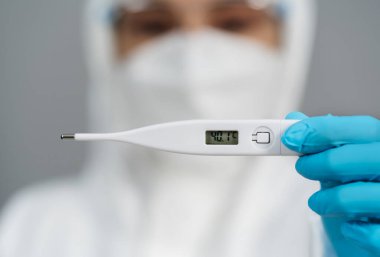 Koruyucu PPE kıyafetli doktor yüksek sıcaklıkta dijital termometre tutuyor.