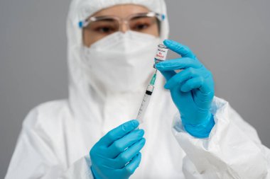 Korumalı PPE kıyafetli doktor Coronavirus (Covid-19) aşı şişesini şırınga ilacına çiziyor