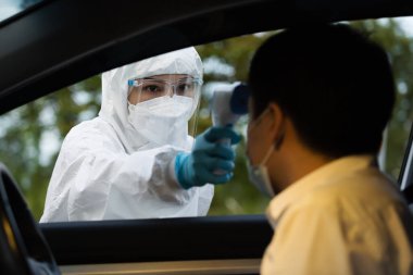 Kızıl ötesi termometre kullanarak koruyucu PPE kıyafetli doktor sürücü istasyonundaki adamın ısısını ölçüyor.