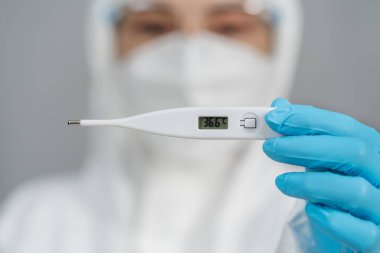 Koruyucu PPE kıyafetli doktor normal sıcaklıkta dijital termometre tutuyor.
