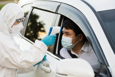Kızıl ötesi termometre kullanarak koruyucu PPE kıyafetli doktor sürücü istasyonundaki adamın ısısını ölçüyor.