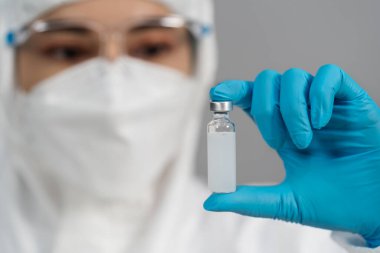 Koruyucu PPE kıyafetli doktor elinde aşı şişesi tutuyor ve nazikçe sallıyor.