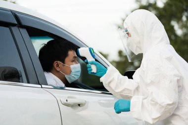 Kızıl ötesi termometre kullanarak koruyucu PPE kıyafetli doktor sürücü istasyonundaki adamın ısısını ölçüyor.