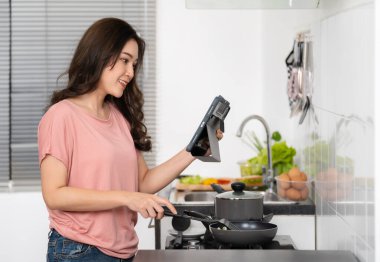 Yemek pişiren ve hazırlayan genç bir kadın evdeki mutfakta bir tablet bilgisayarın tarifine göre...