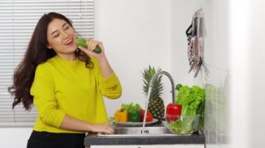 Salatalıkla şarkı söyleyen ve evdeki mutfaktaki lavaboda sebze yıkayan mutlu genç kadın.